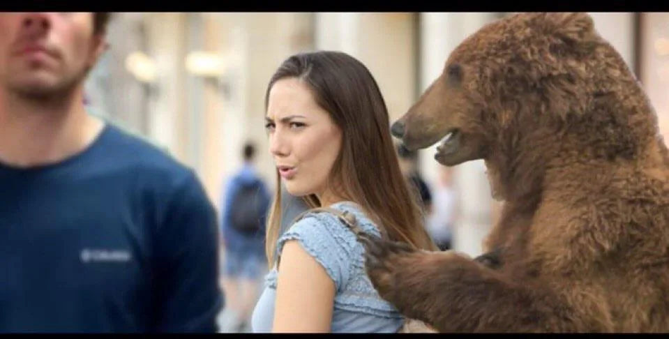 A woman in a blue top looks over her shoulder with a confused and annoyed expression as a large brown bear stands beside…