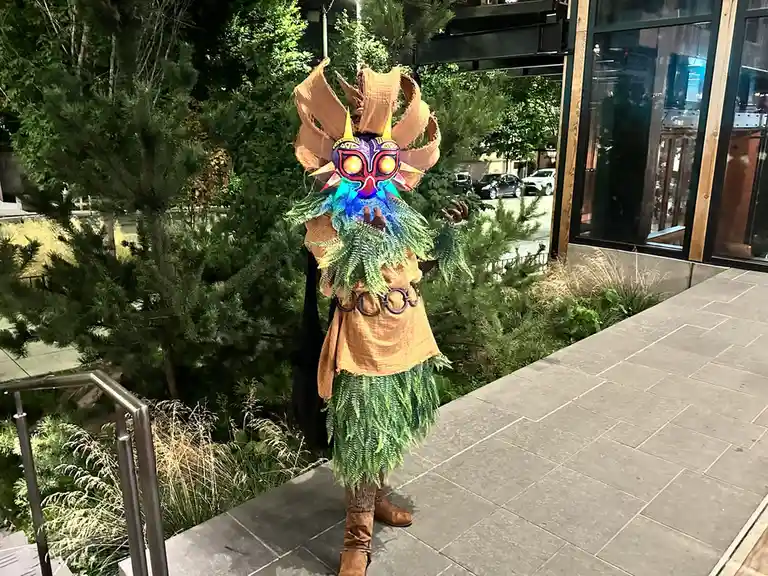 A person in a detailed costume resembling the video game character Majora's Mask stands outdoors at night, wearing a glowing…
