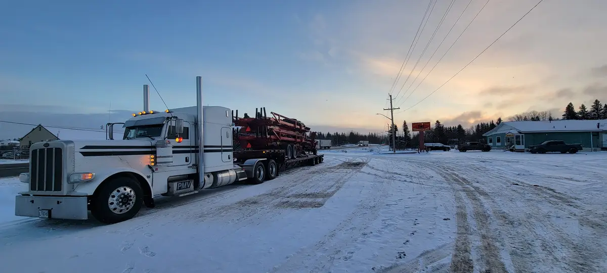 A large white semi-truck with a red flatbed trailer is parked on a snow-covered road at dusk, with tire tracks and…
