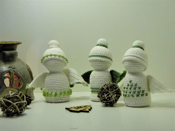 Three crocheted angel figurines with white bodies and green accents stand on a white surface next to decorative items. The…