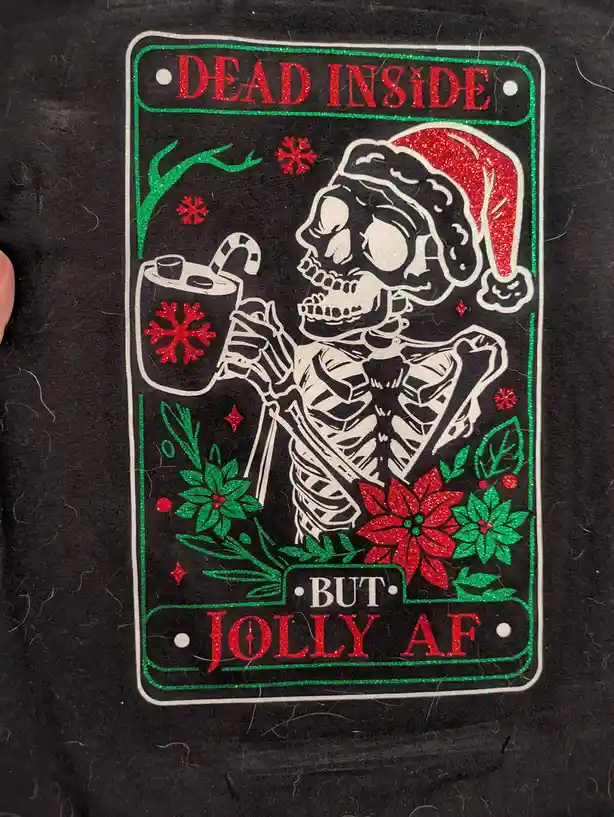 A glittery black t-shirt graphic shows a skeleton wearing a red Santa hat, holding a mug with a candy cane stirrer and a red…