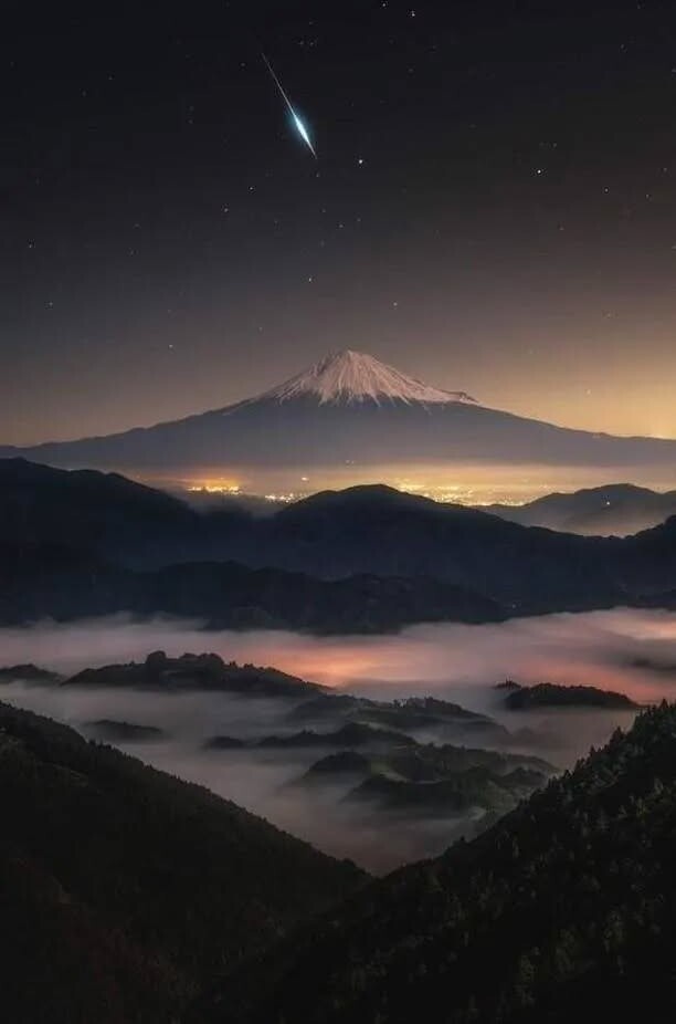 A breathtaking night photograph of Mount Fuji, its snow-capped peak silhouetted against a starry sky with a bright shooting…