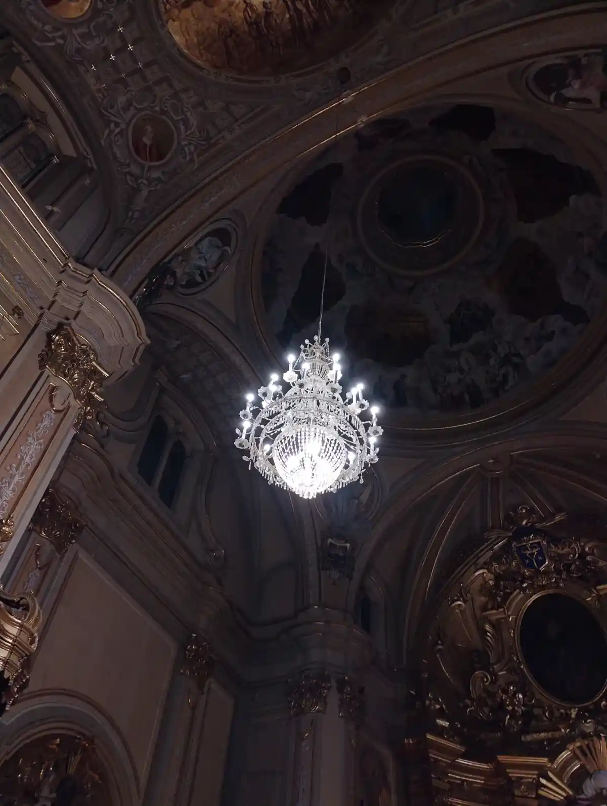 A photo looking up at an ornate, baroque-style church ceiling with frescoes, gold trim, and a large, bright crystal…