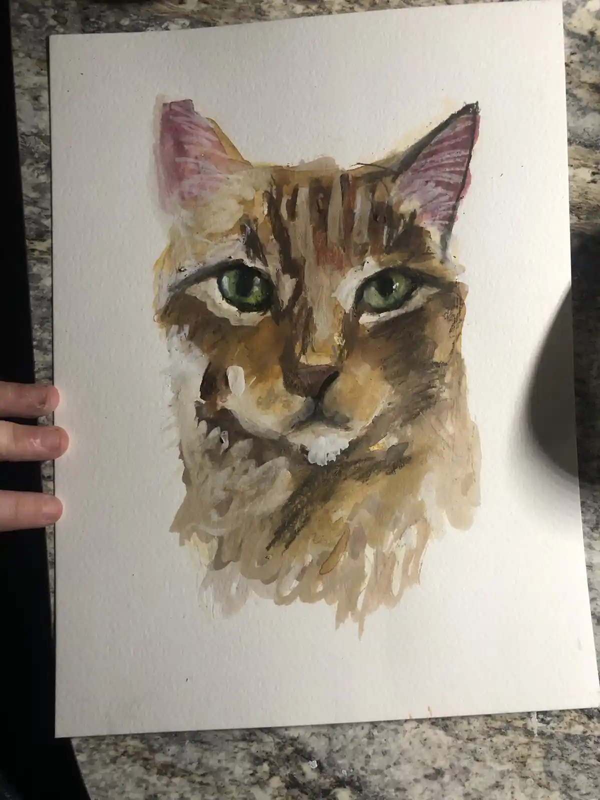 A watercolor painting of a cat's face with green eyes and brown fur, held by a hand on a speckled countertop. The overlaid…