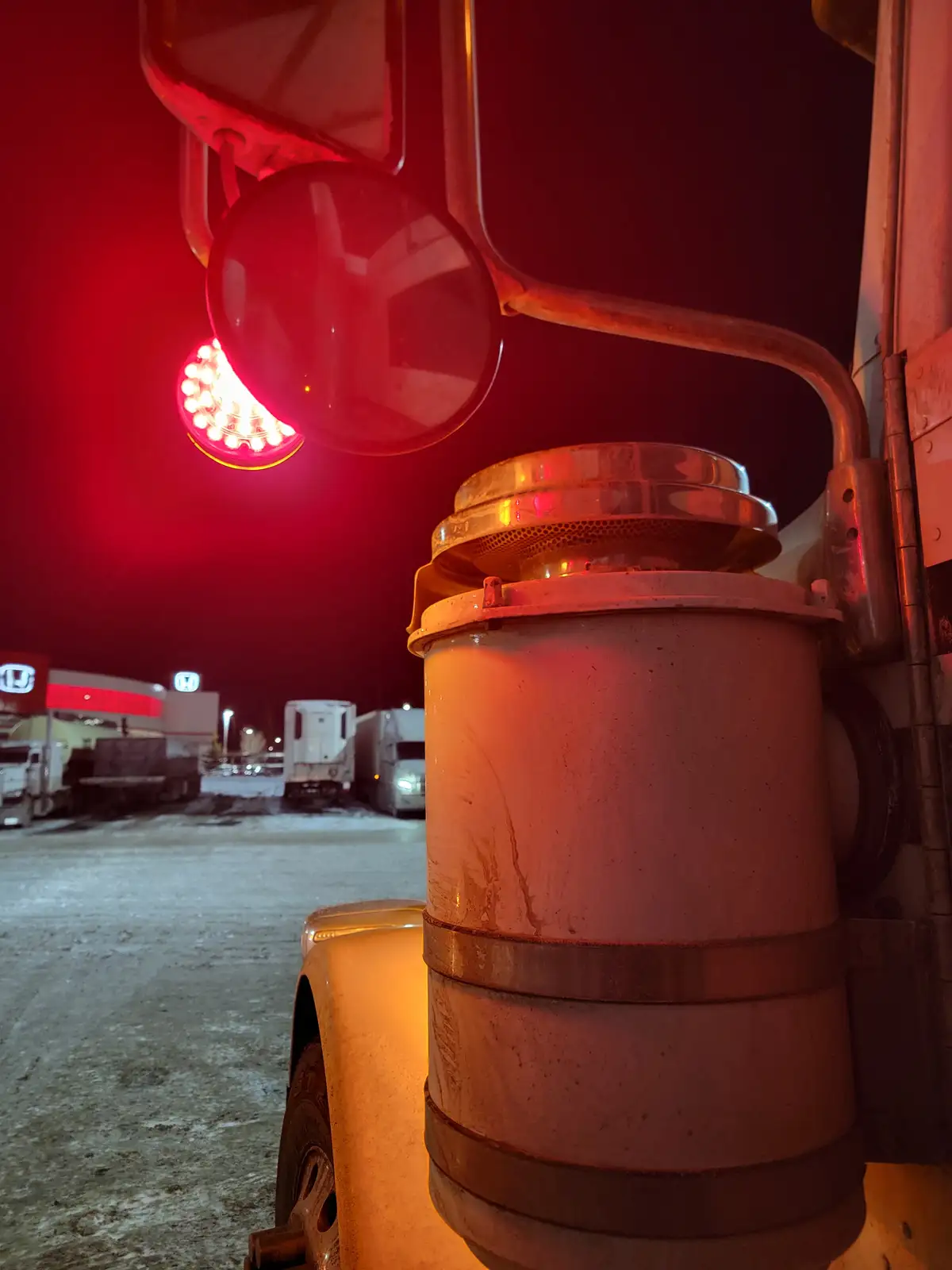 A close-up nighttime photo of a large truck's side mirror and a cylindrical light housing, both bathed in red light. The…