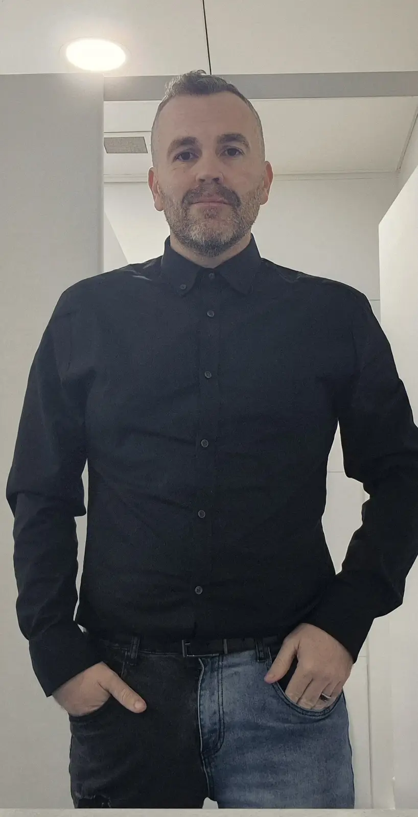 a man with a beard and short hair stands facing the camera, wearing a black button-up shirt and jeans that are split down…