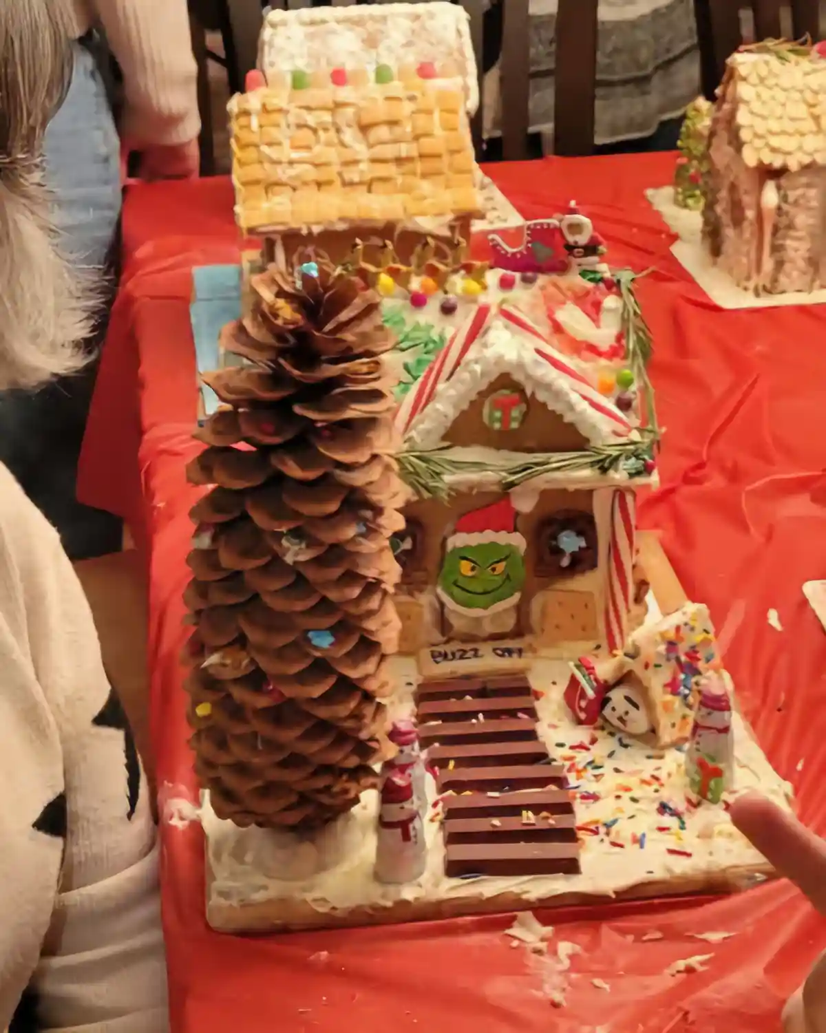 A gingerbread house decorated with a giant pinecone as a tree, a Grinch face on the door, candy canes, and a sign that reads…