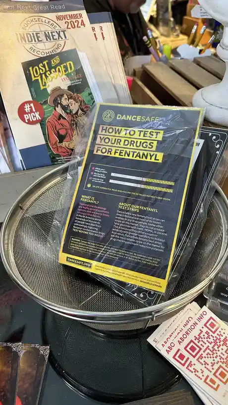 A DanceSafe fentanyl test strip instruction pamphlet sits in a metal strainer on a table, next to a book titled 'Lost and…