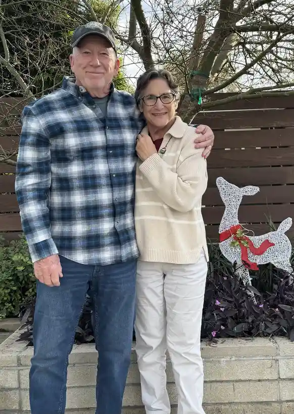 An older couple poses for a photo outdoors, smiling and embracing. The man wears a plaid shirt and cap, the woman wears…