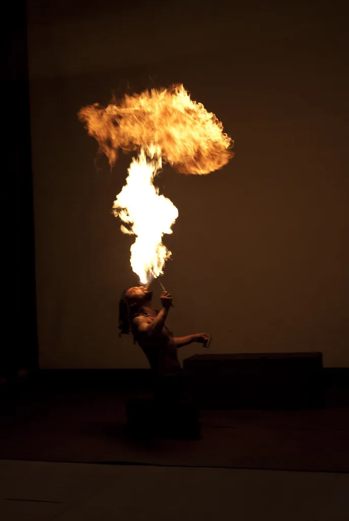 A person is kneeling and blowing fire from their mouth, creating a large, bright flame that rises upward. The person is…