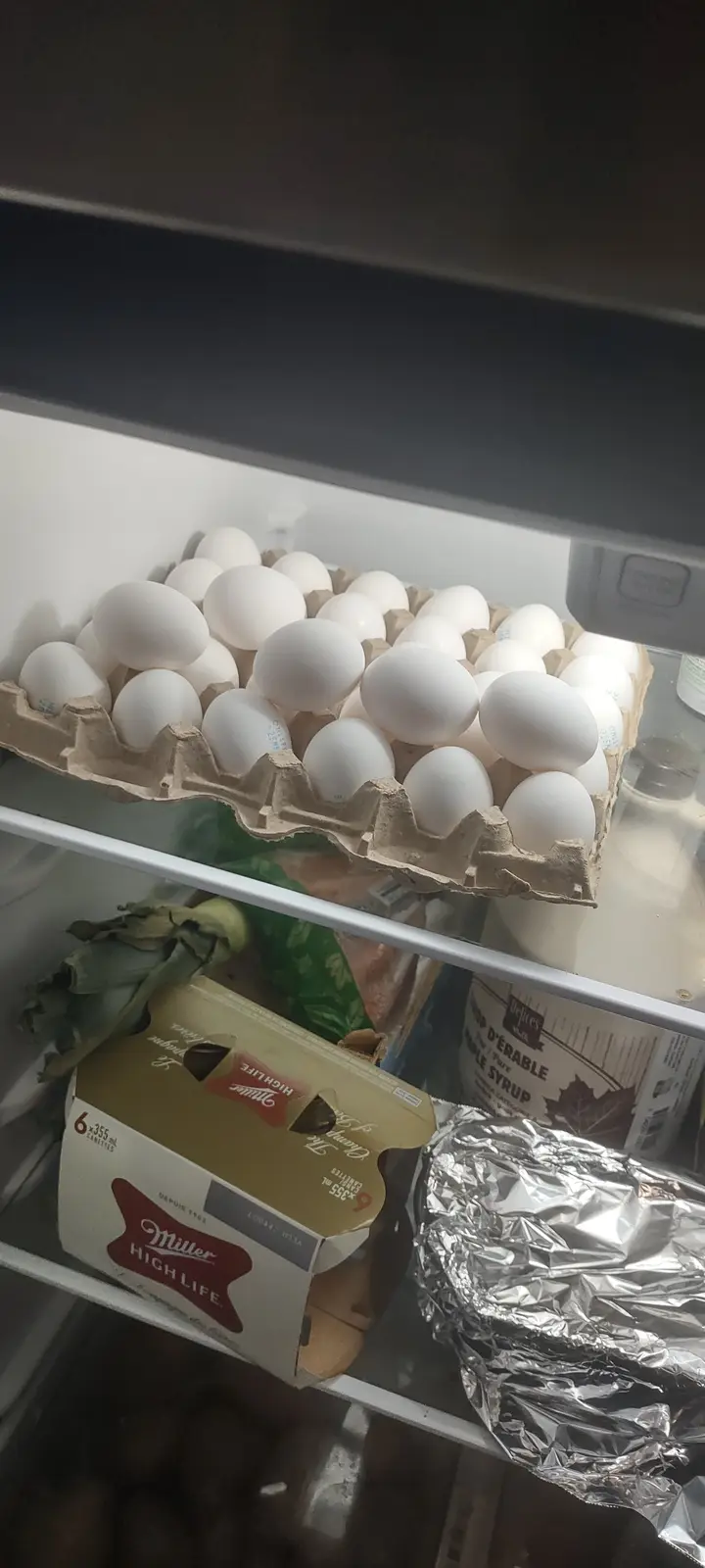 A refrigerator shelf holds a large carton of white eggs on the top shelf, with a Miller High Life beer six-pack and…