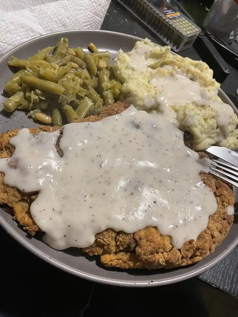 A close-up photo of a plate of chicken fried steak smothered in white gravy, accompanied by green beans and mashed potatoes…
