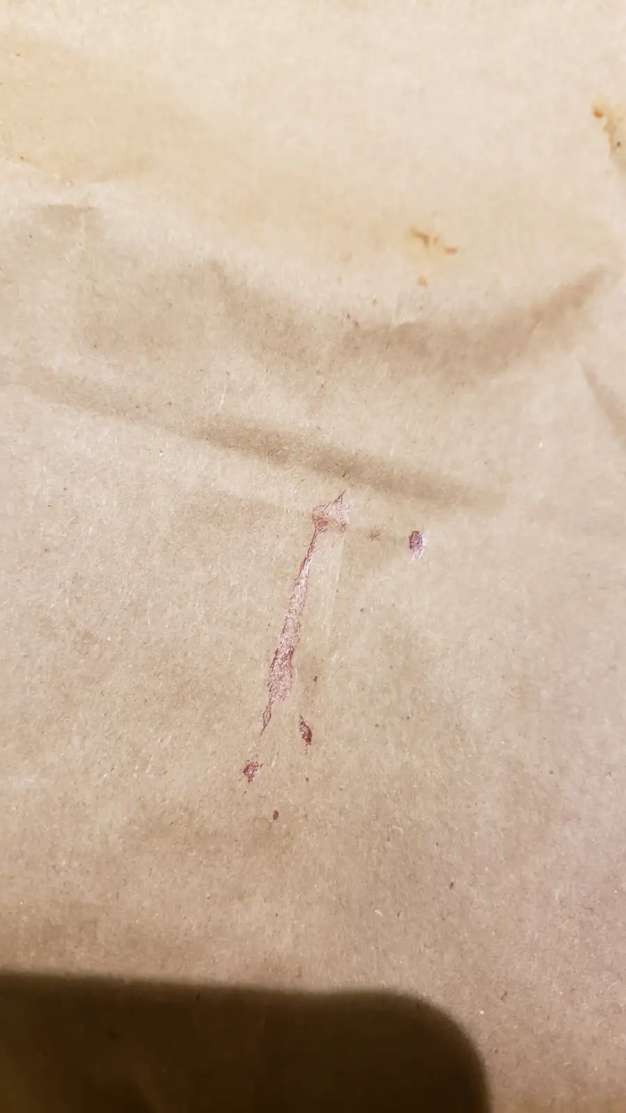 A close-up photo shows a brown paper surface with a faint, reddish-brown stain that resembles a single drop of blood with a…