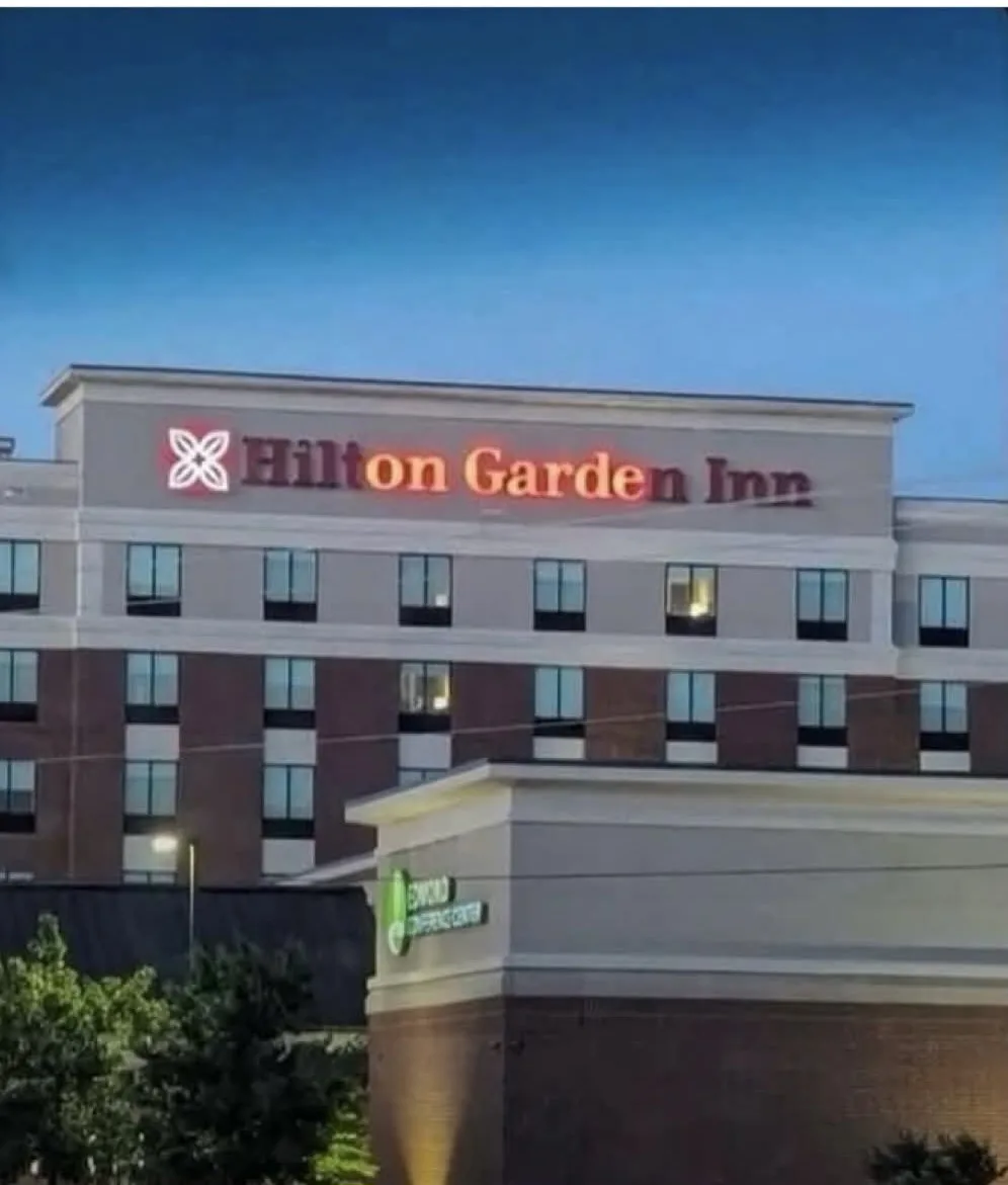 An exterior shot of a Hilton Garden Inn hotel building at dusk, with its illuminated sign visible against a clear blue sky…