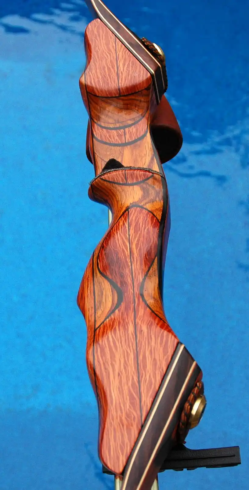 A close-up shot of a highly ornate, custom wooden bow with dark wood grain and black inlaid lines, set against a blue…