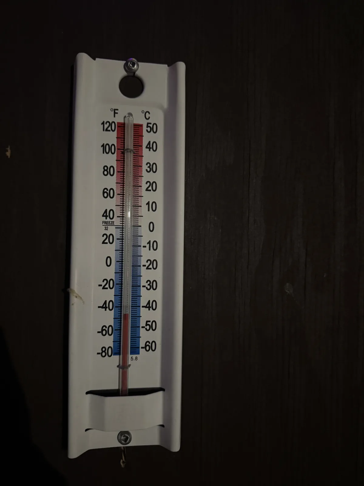 A white outdoor thermometer mounted on a dark wooden surface shows a temperature of 5.8 degrees Celsius (42.4 degrees…