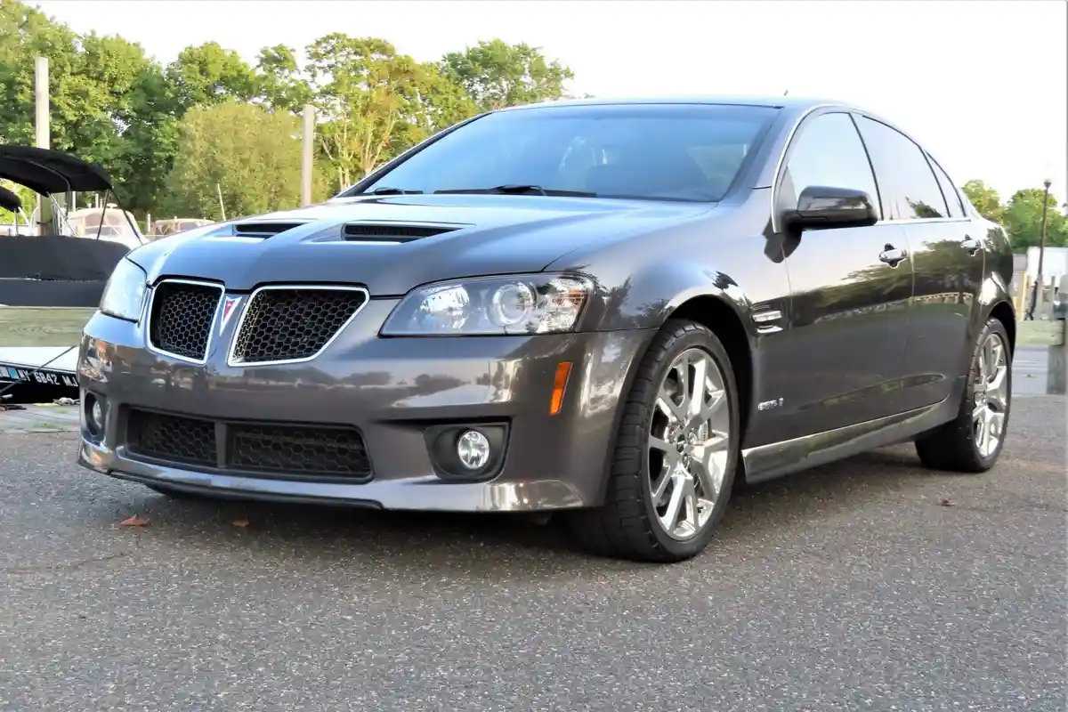 A dark gray Pontiac G8 GXP sedan is parked on asphalt with trees and a boat in the background. The car has a prominent front…