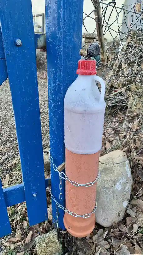 A white plastic water bottle with a red cap, partially frozen and covered in frost, is chained to a blue wooden fence post…