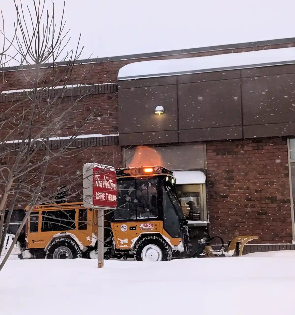 A yellow snowplow truck is parked at a Tim Hortons drive-thru during a snowstorm, with snow falling and covering the ground…