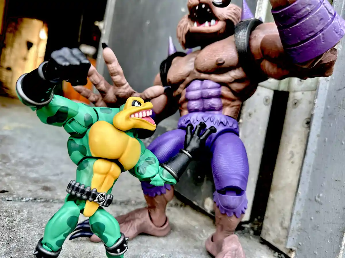 A green and yellow reptilian action figure with a wide grin and black-gloved fists stands defiantly in front of a larger…