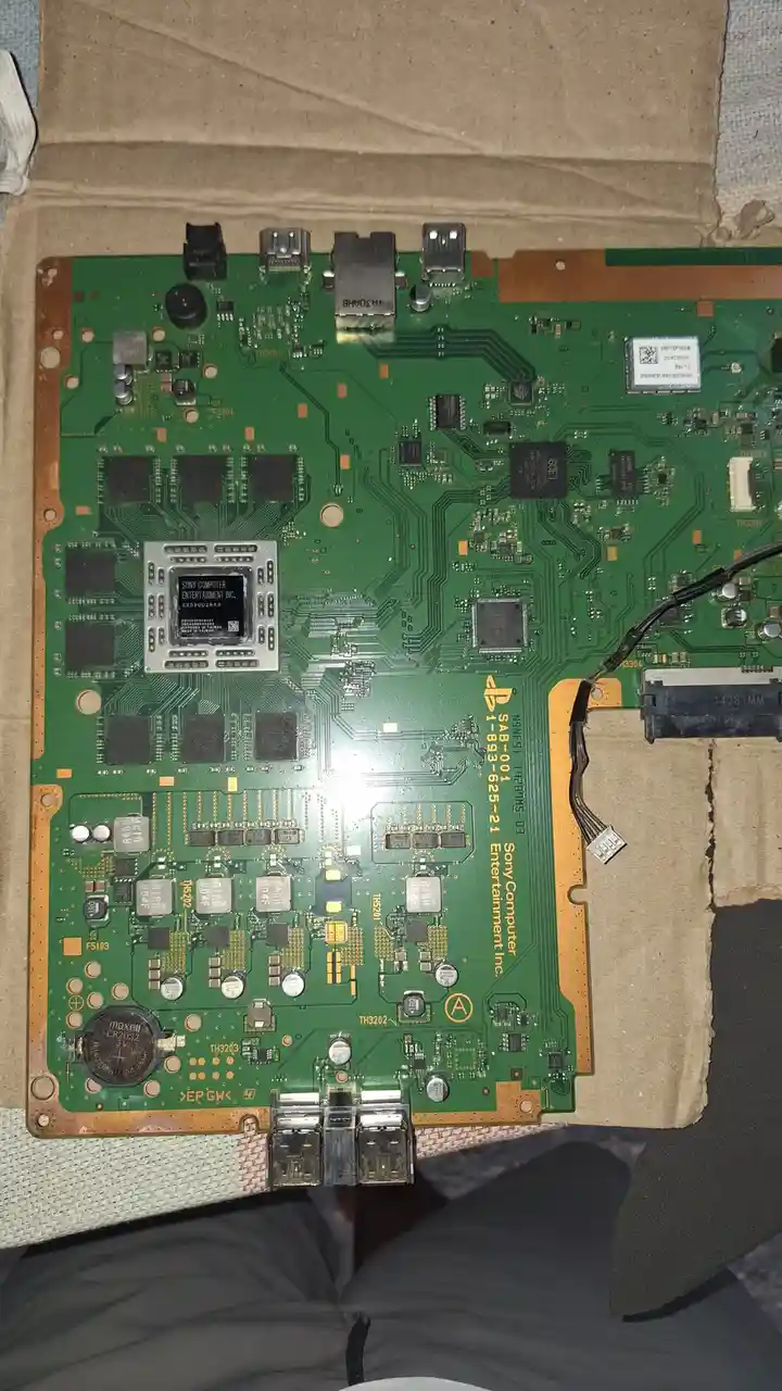 A top-down photo of a green PlayStation 5 motherboard with visible circuitry, chips, and ports, resting on cardboard. The…