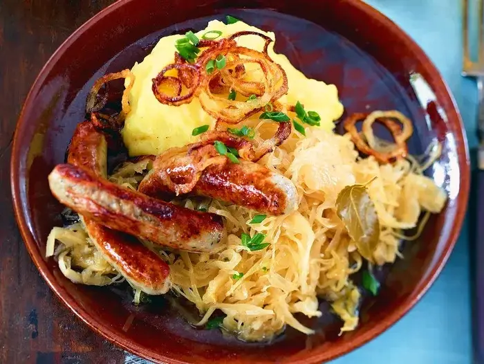 A close-up photo of a bowl of food containing sauerkraut, several grilled sausages, mashed potatoes, and crispy fried…