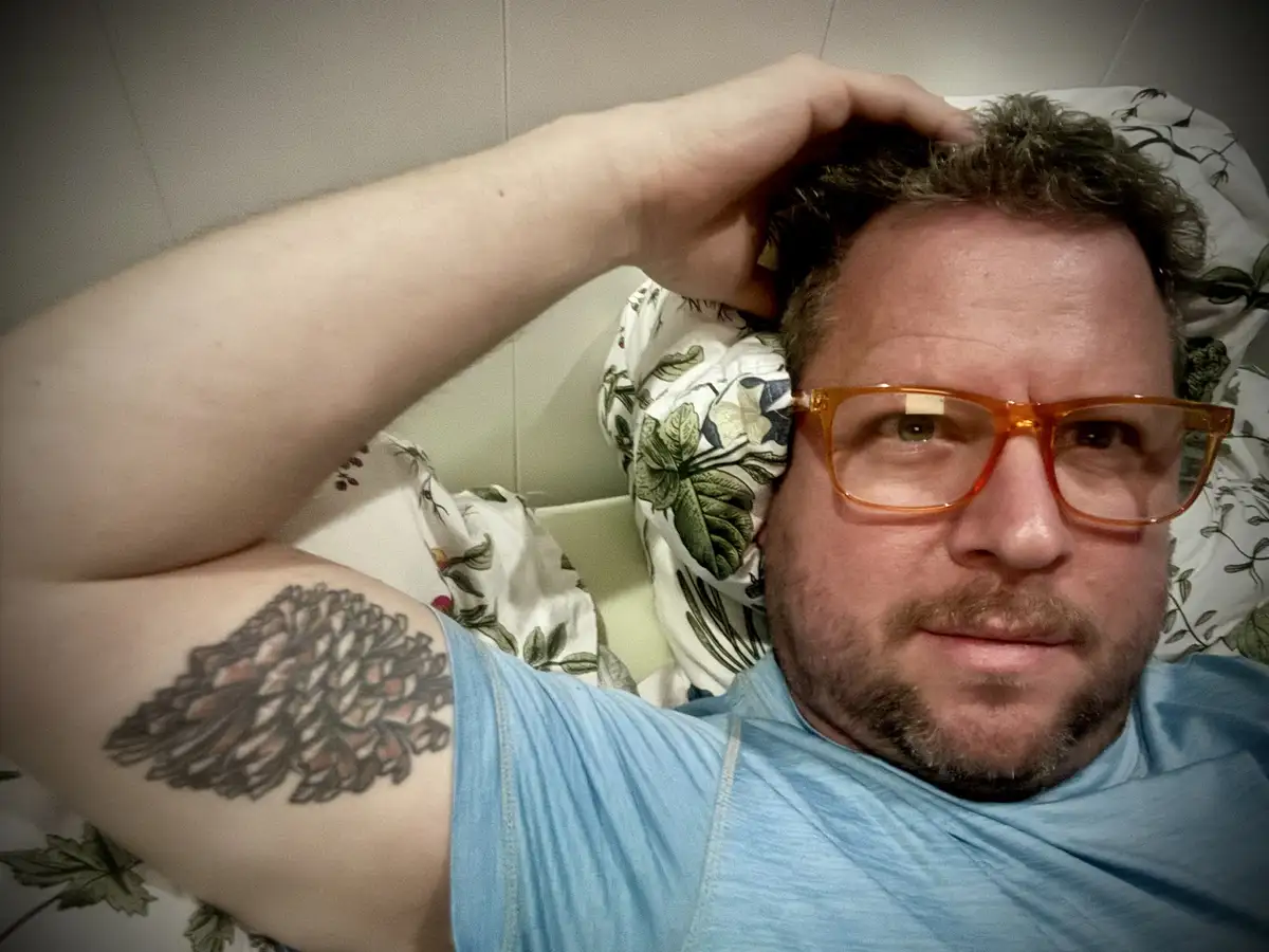A man with curly hair and orange glasses lies in bed, his arm raised to his head, revealing a detailed pinecone tattoo on…