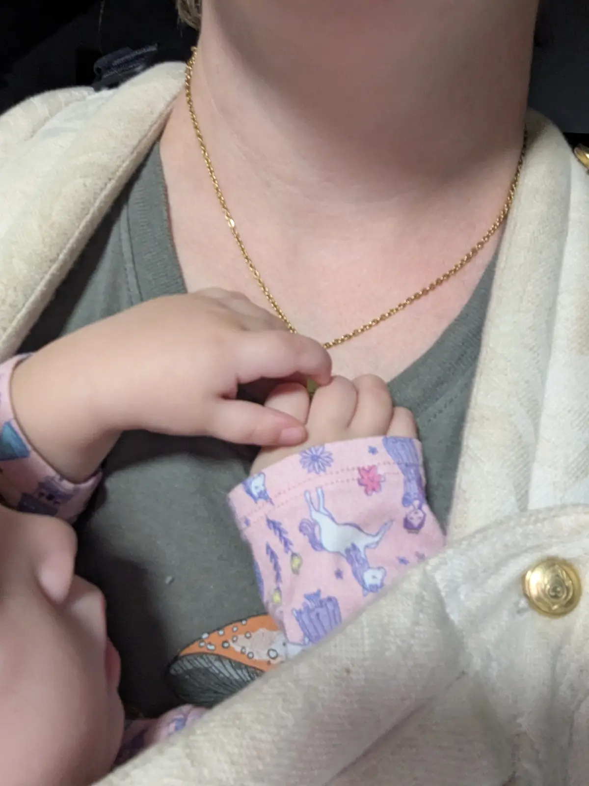A close-up photo shows a toddler's hand gripping an adult's gold chain necklace, while the adult wears a beige coat over a…