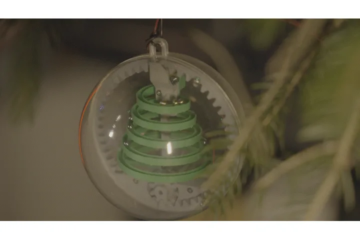 A close-up of a mechanical Christmas tree ornament with green spiral tiers inside a clear glass ball, hanging on a pine…