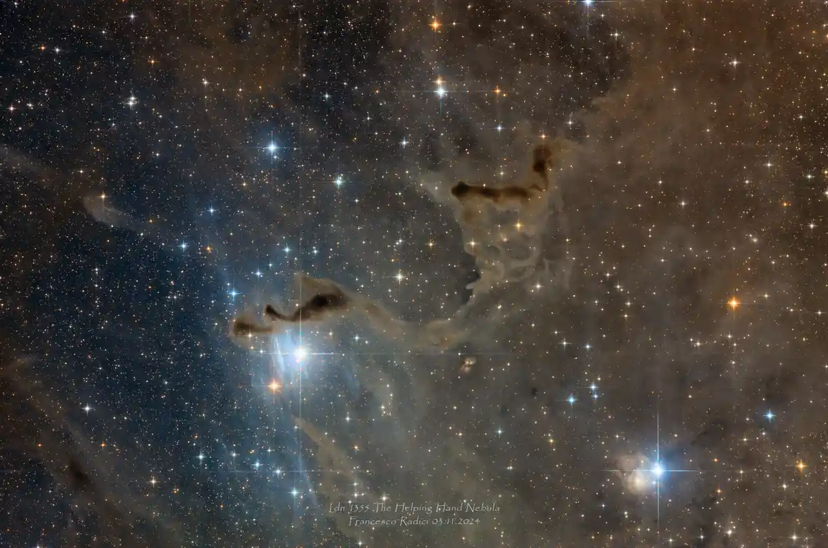 A deep-space photograph of the Ldn 1355 nebula, also known as the Helping Hand Nebula, showing a dark, finger-like cloud…
