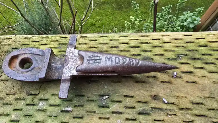 A rusty, industrial-looking metal tool with a pointed tip and a looped handle rests on a weathered, perforated wooden…
