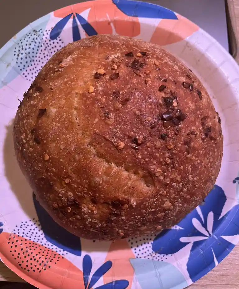 A round, golden-brown loaf of bread with visible seeds and a crack on top sits on a colorful paper plate with blue, orange…