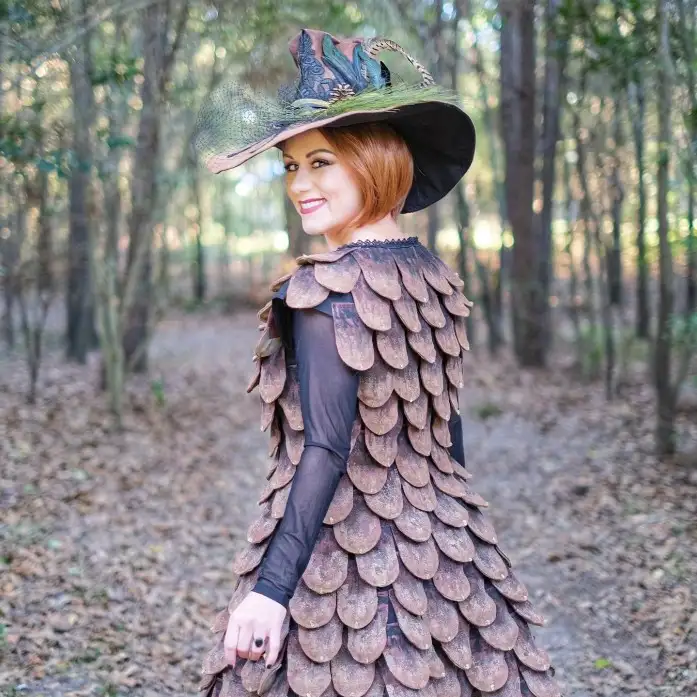 A woman with short red hair stands in a forest, wearing a large ornate hat and a dress made of overlapping brown wooden…