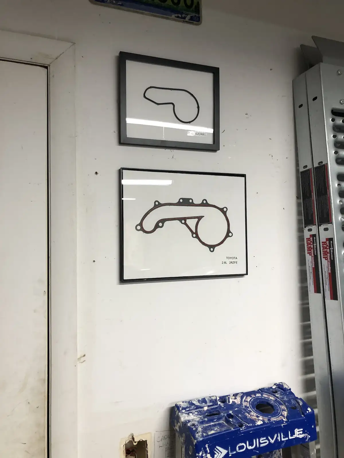Two framed automotive gasket outlines hang on a white wall in what appears to be a garage or workshop. The top frame…