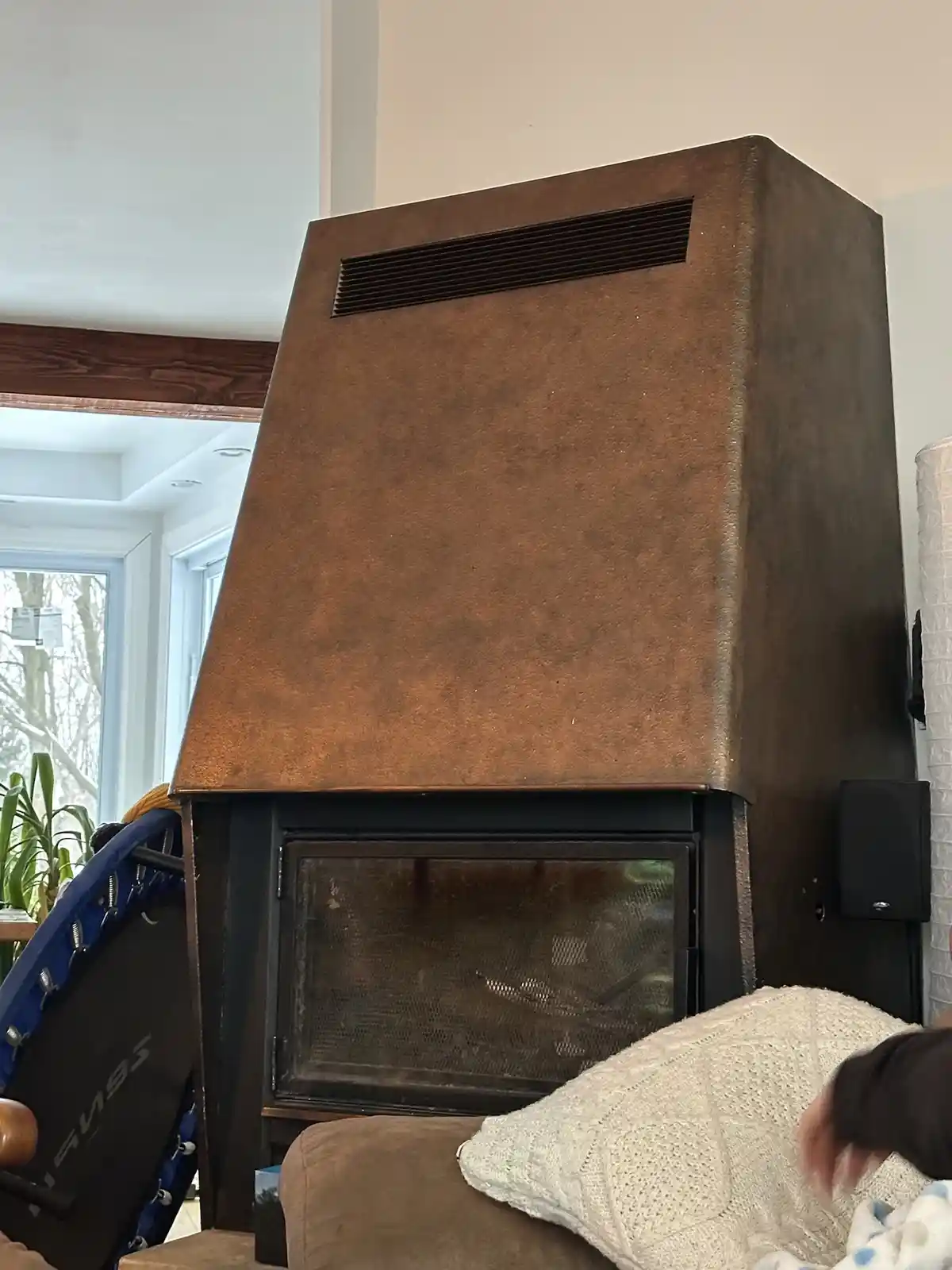 A large, brown, boxy fireplace with a vent on top stands in a living room, next to a couch with a white blanket and a blue…