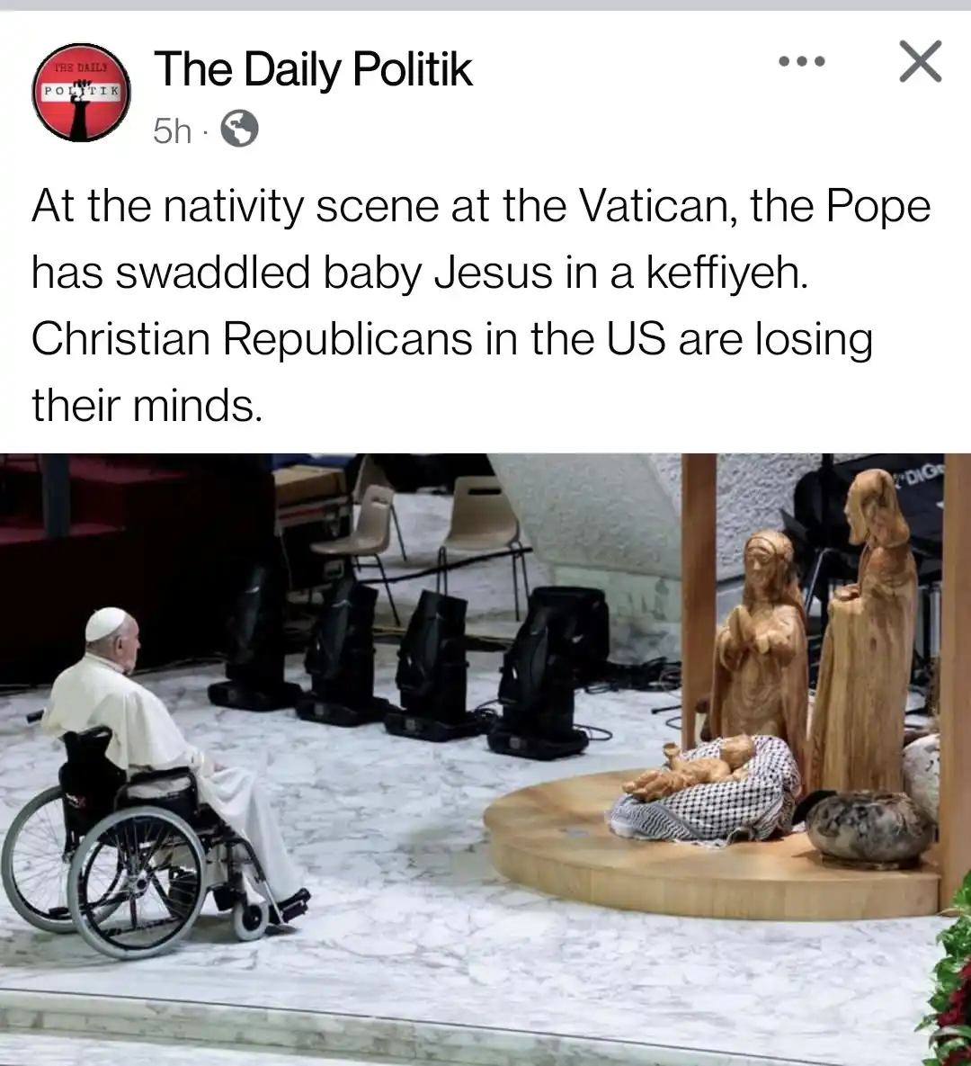 A photograph shows Pope Francis in a wheelchair, looking toward a wooden nativity scene. In the scene, baby Jesus is…