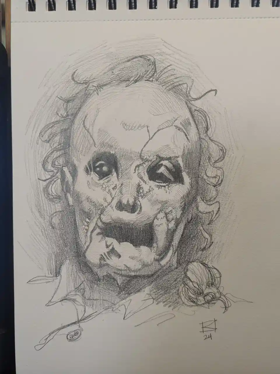 A pencil sketch on spiral-bound paper depicts a grotesque, skeletal face with wild hair, stitched skin, and a wide, toothy…