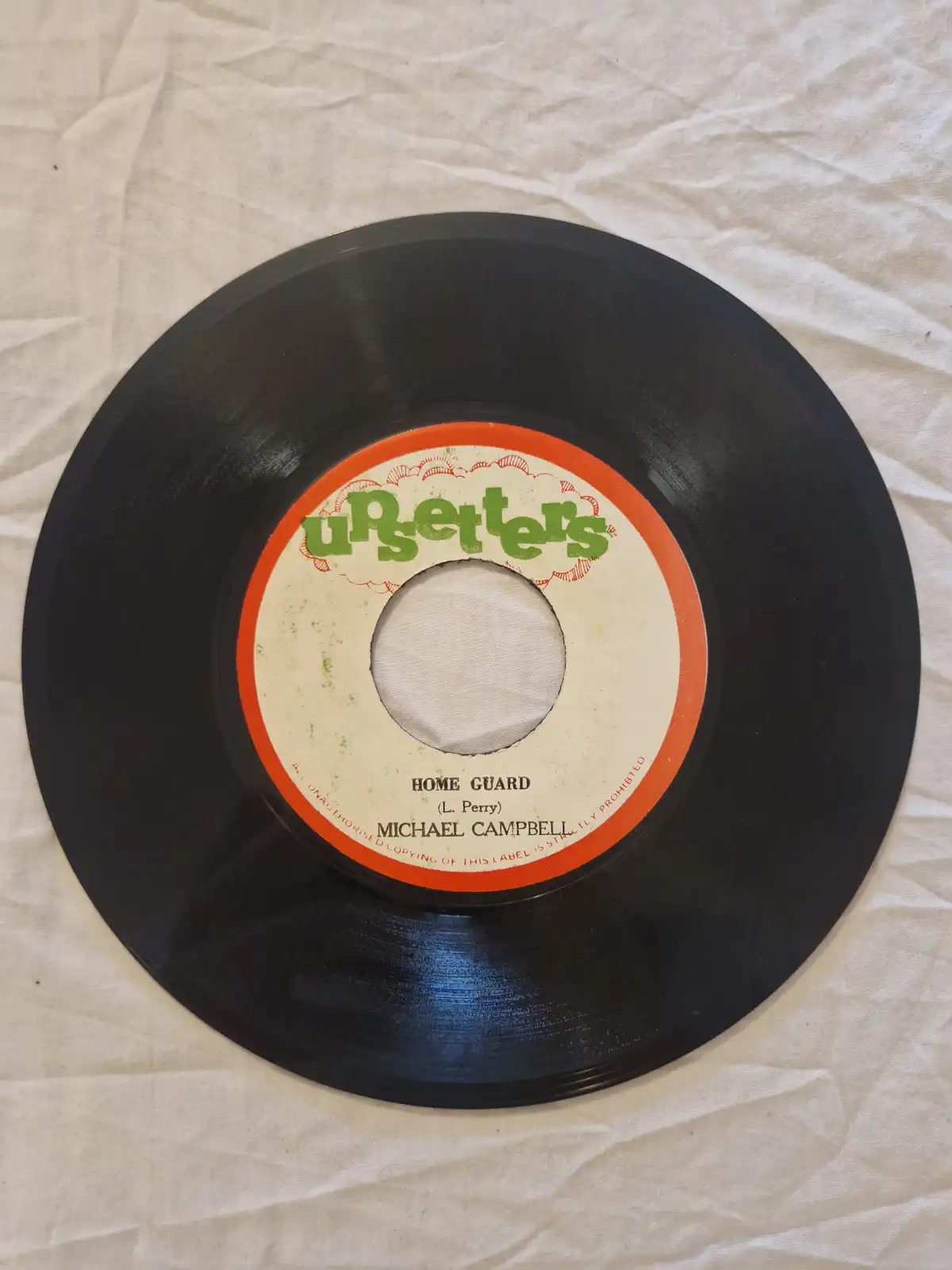 A close-up photo of a black 7-inch vinyl record resting on a wrinkled white fabric surface. The record's label is…