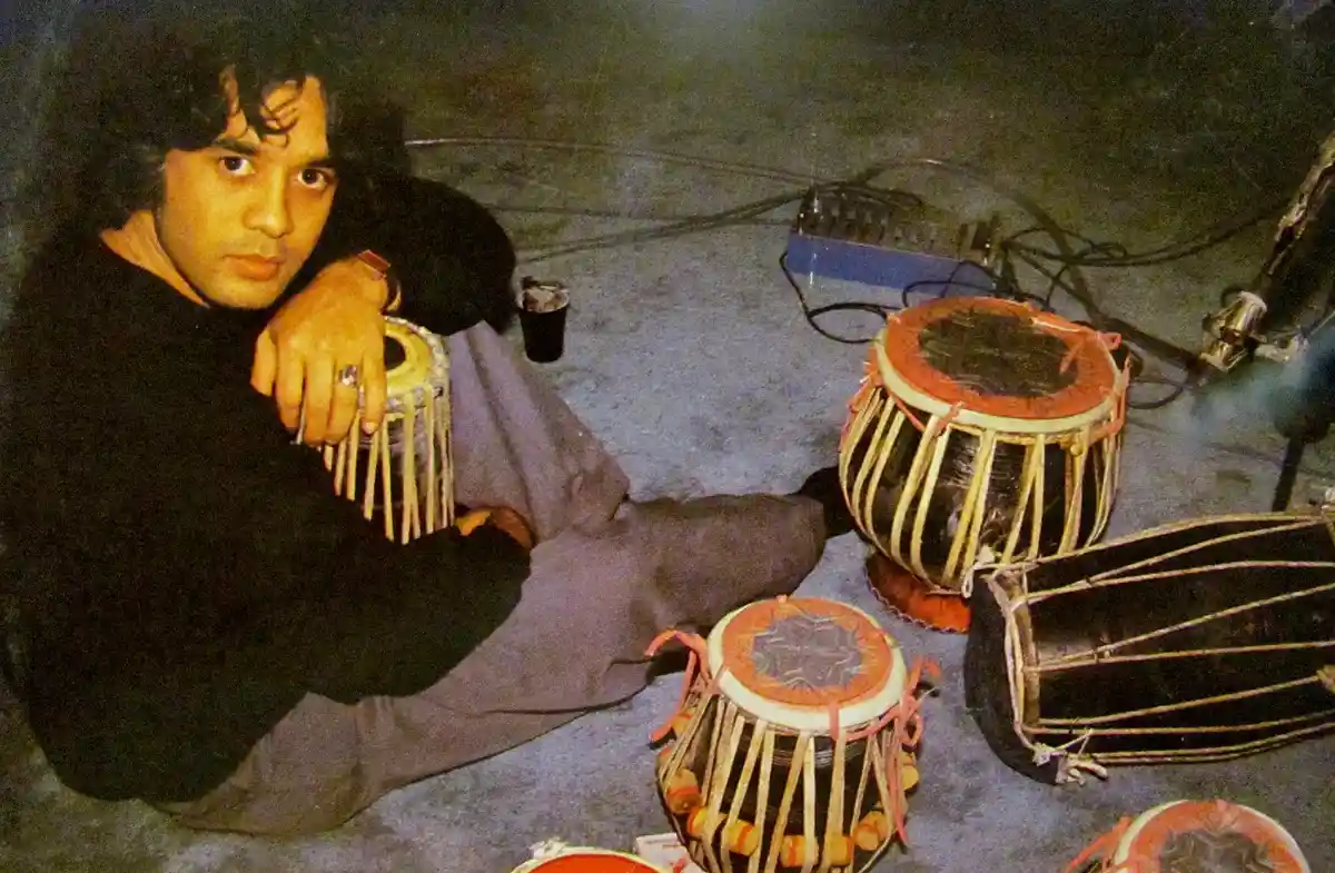 a man with dark curly hair sits cross-legged on a carpeted floor, holding a small tabla drum, surrounded by several other…