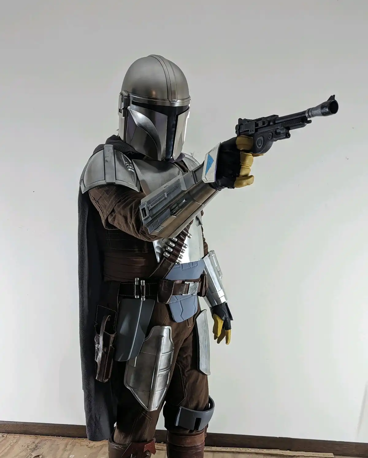 A person in a detailed Mandalorian cosplayer costume, featuring a silver helmet, brown armor, and a black cape, stands…