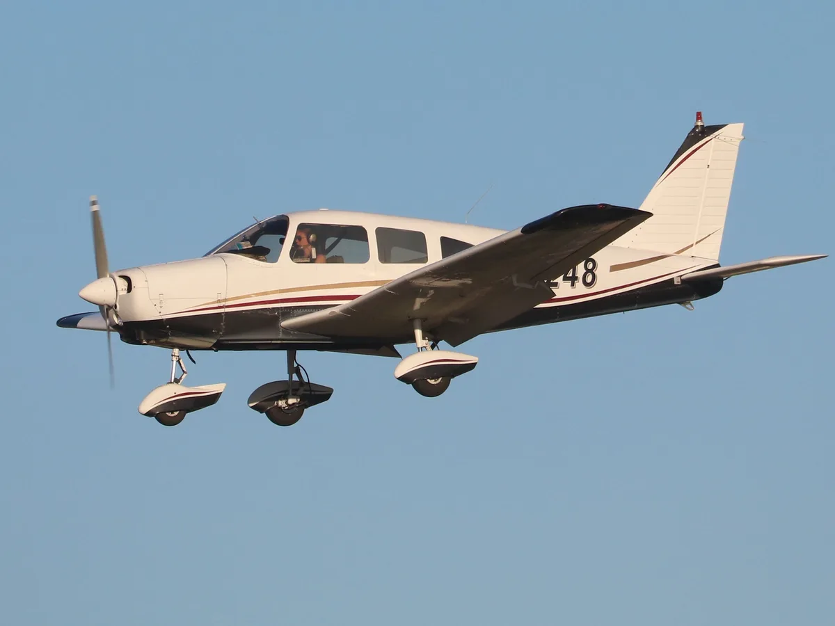A white single-engine propeller airplane with black and gold stripes is flying low against a clear blue sky, its landing…