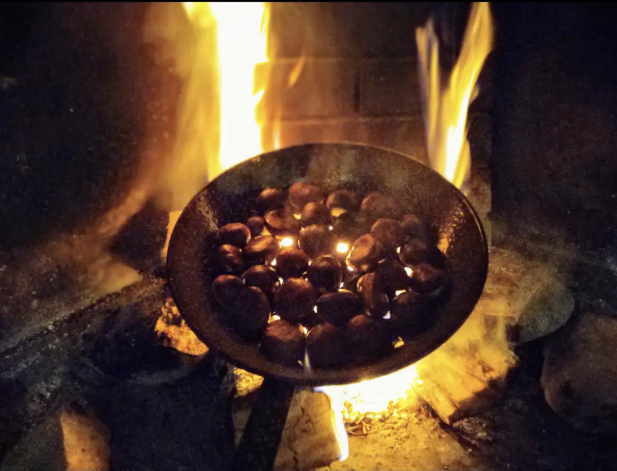 A dark metal pan filled with chestnuts sits over an open fire, with flames licking up around it and glowing embers visible…