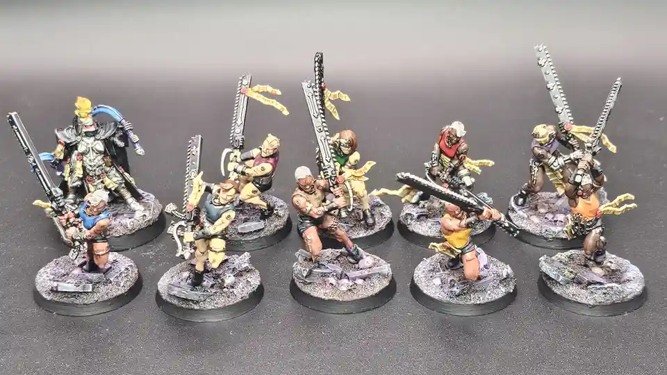 Seven painted miniature figurines, resembling grimdark fantasy warriors with chainswords and armor, are arranged in a row on…