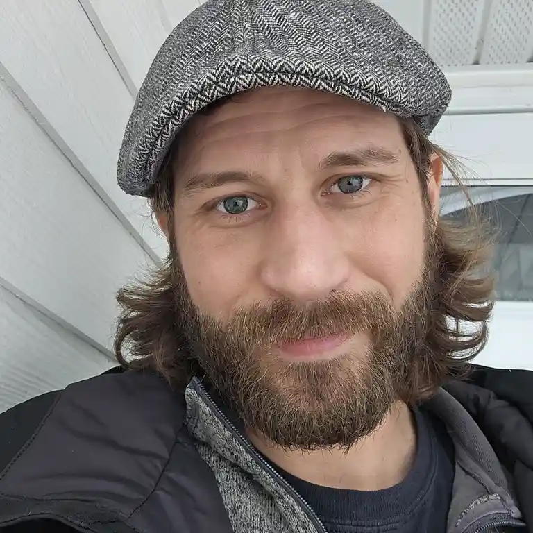 A close-up selfie of a man with long brown hair, a full beard, and a grey herringbone flat cap, looking directly at the…