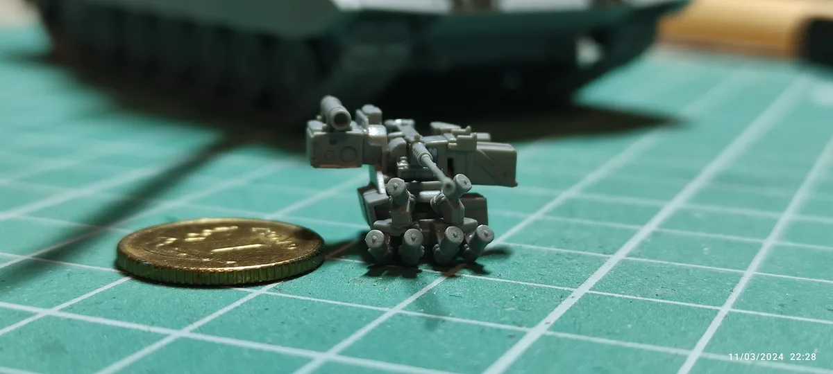 A small, grey plastic model of a multi-barrelled weapon system sits on a green gridded cutting mat next to a gold-colored…