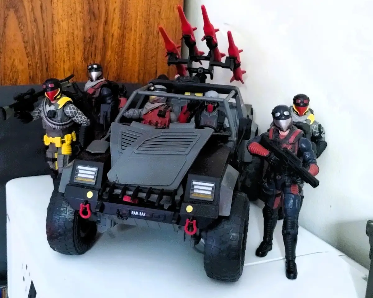 A toy military vehicle with multiple armed action figures posed around it, including some with red masks and yellow accents…