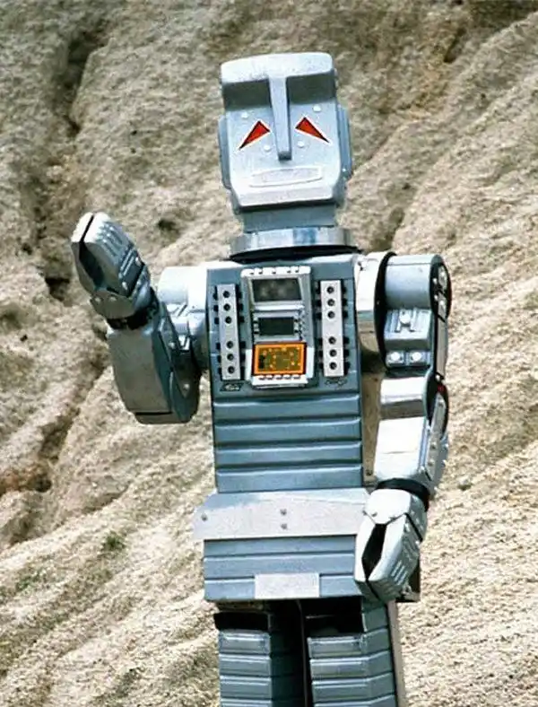 A vintage silver robot with red triangular eyes and a digital display on its chest stands outdoors against a rocky…