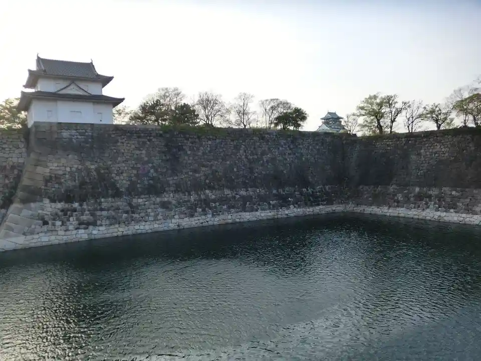 A wide shot of a traditional Japanese castle with a large stone wall and moat filled with dark water in the foreground. A…