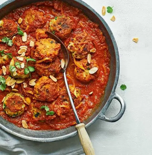 a top-down photo of a metal pan filled with meatballs in a red tomato sauce, garnished with sliced almonds and fresh parsley…