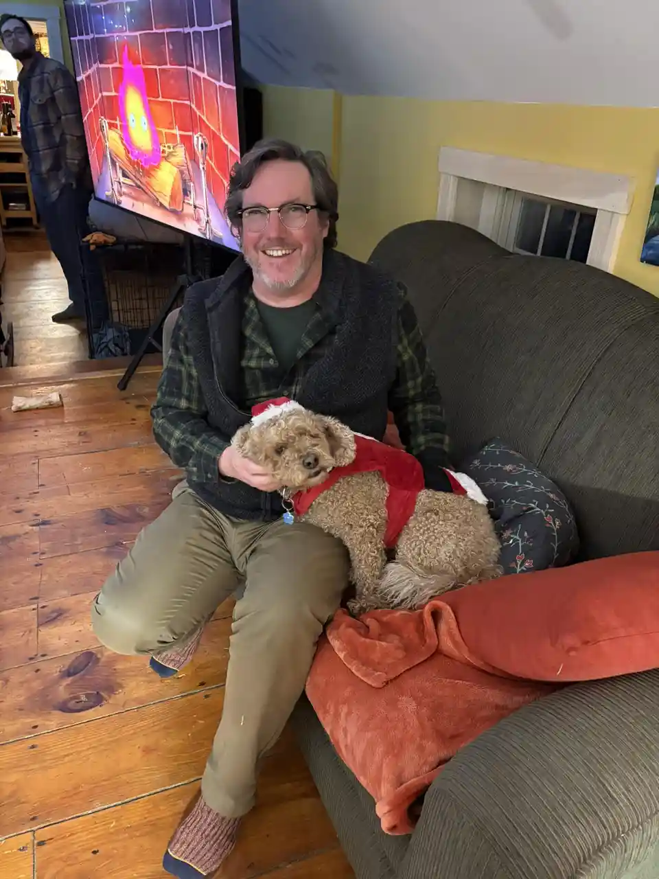 A man with glasses and a beard sits on a couch holding a curly-haired dog wearing a red Santa-style vest. Behind him, a TV…